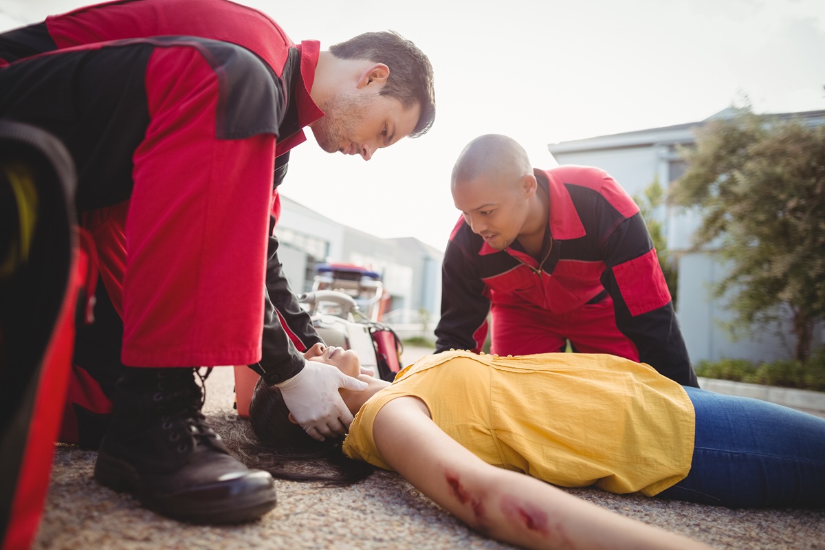 Two paramedics in red uniforms kneeling over an unconscious woman on the ground, one checking her airway.