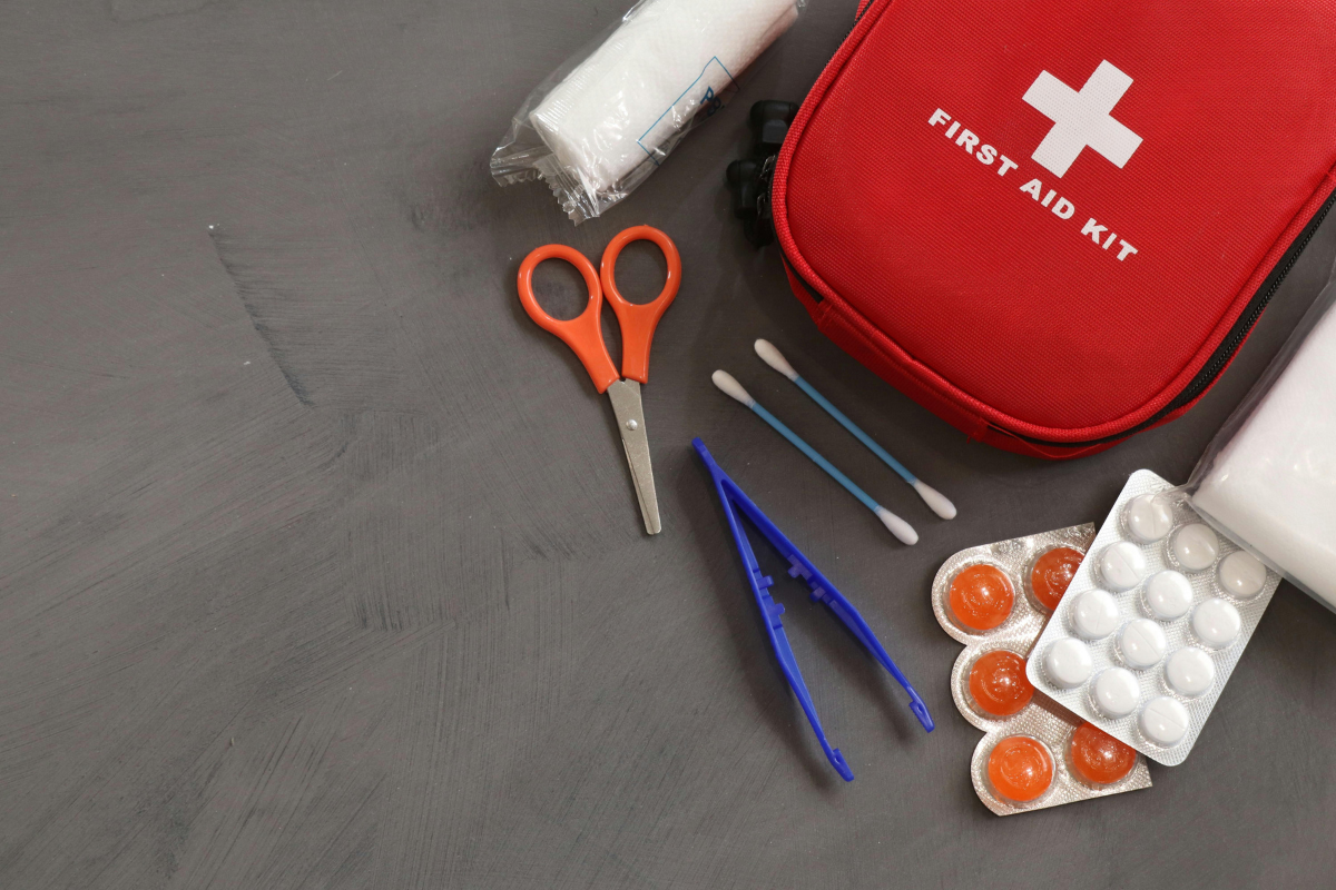 Red first aid kit with scissors, tweezers, gauze, and blister packs on a grey surface