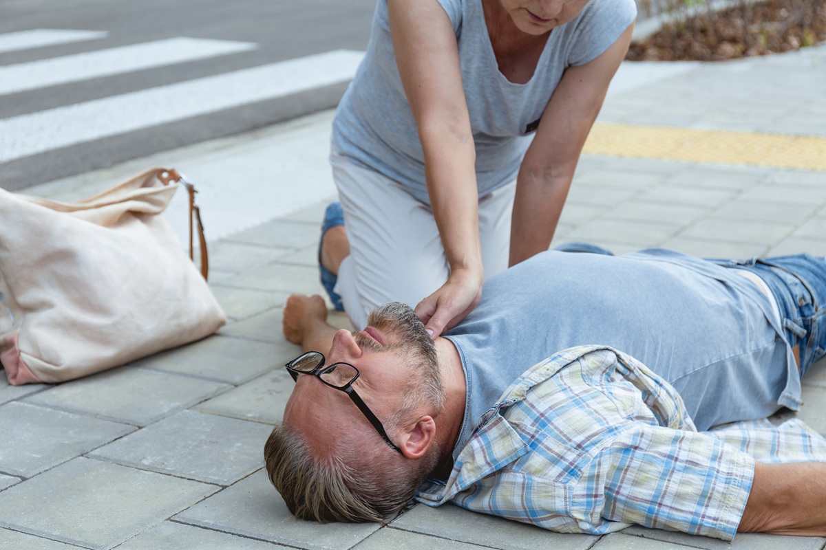 Woman kneeling on a footpath beside an unconscious man, placing a hand on his face to check for a response.