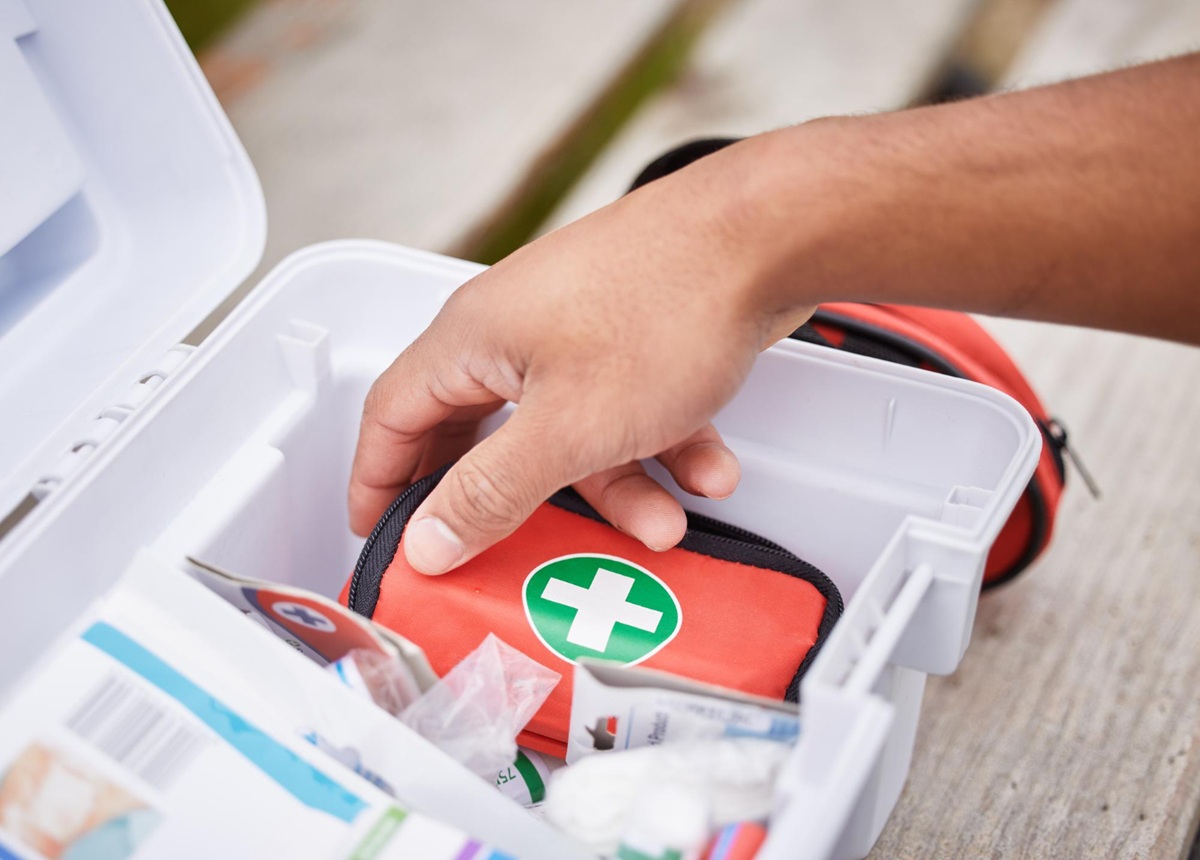 Close-up of a person reaching into a white first aid kit, selecting a red zippered pouch marked with a green cross, surrounded by various medical supplies.