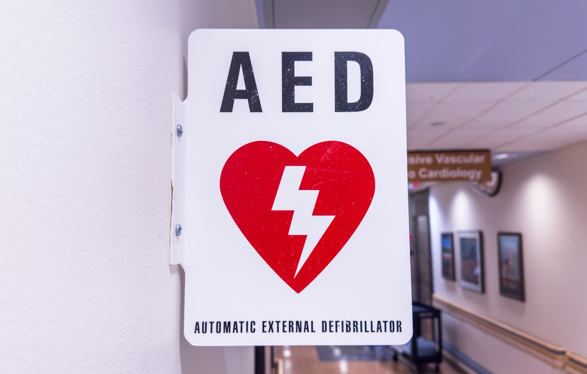 Wall-mounted AED directional sign in a healthcare facility corridor indicating defibrillator location.