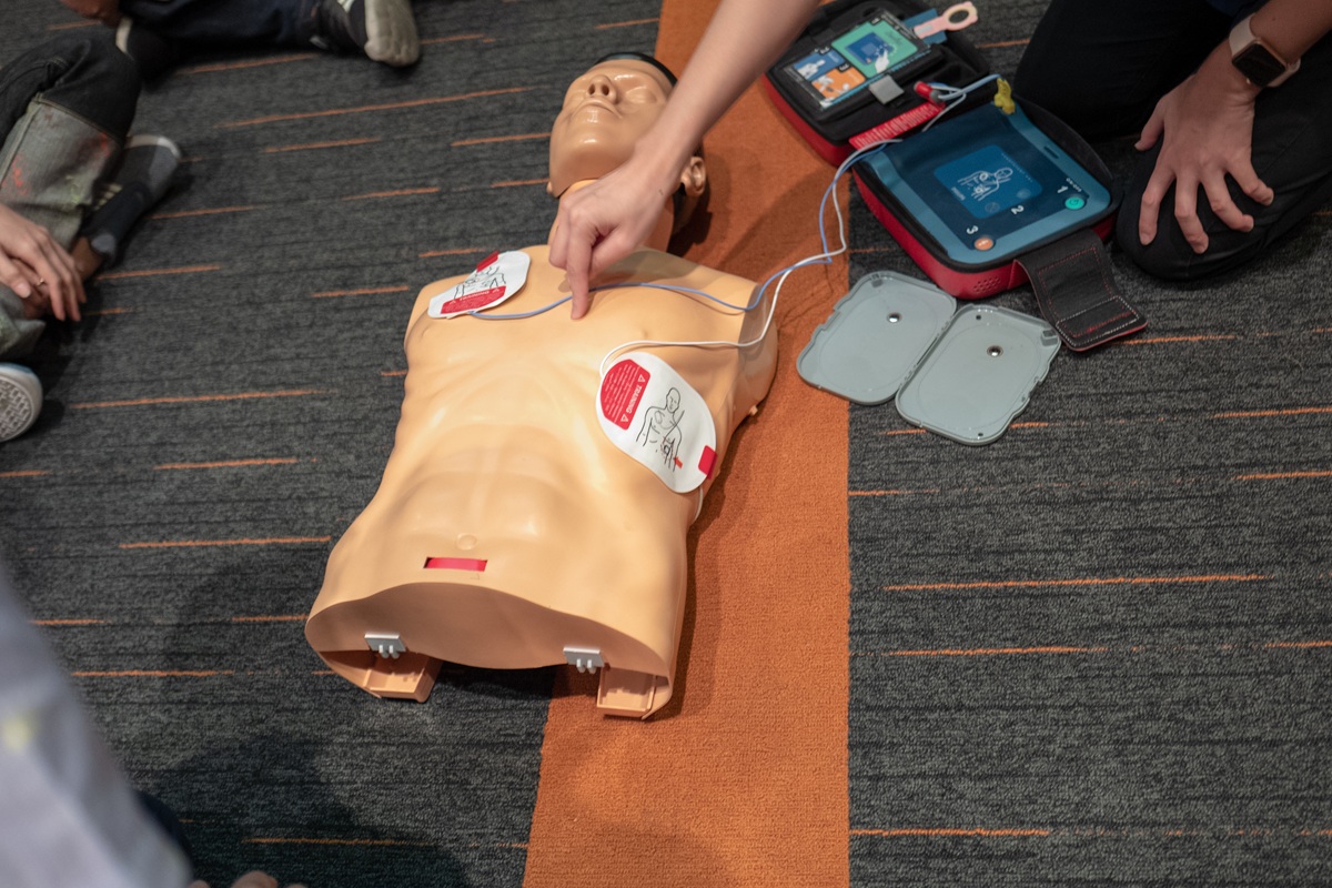 CPR and AED training session using a torso mannequin and defibrillator training pads on the floor.