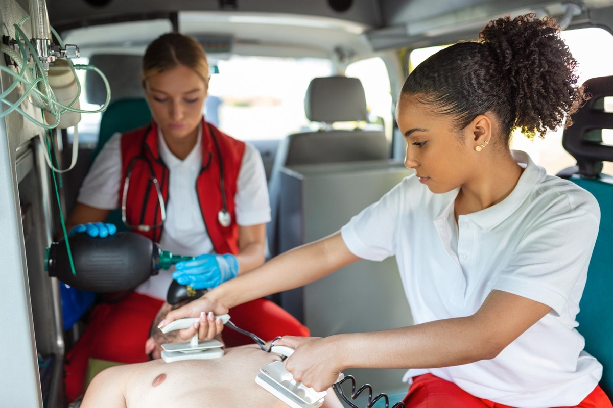 Two paramedics performing emergency care inside an ambulance—one using a defibrillator and the other assisting with ventilation using a bag valve mask on an unconscious patient.