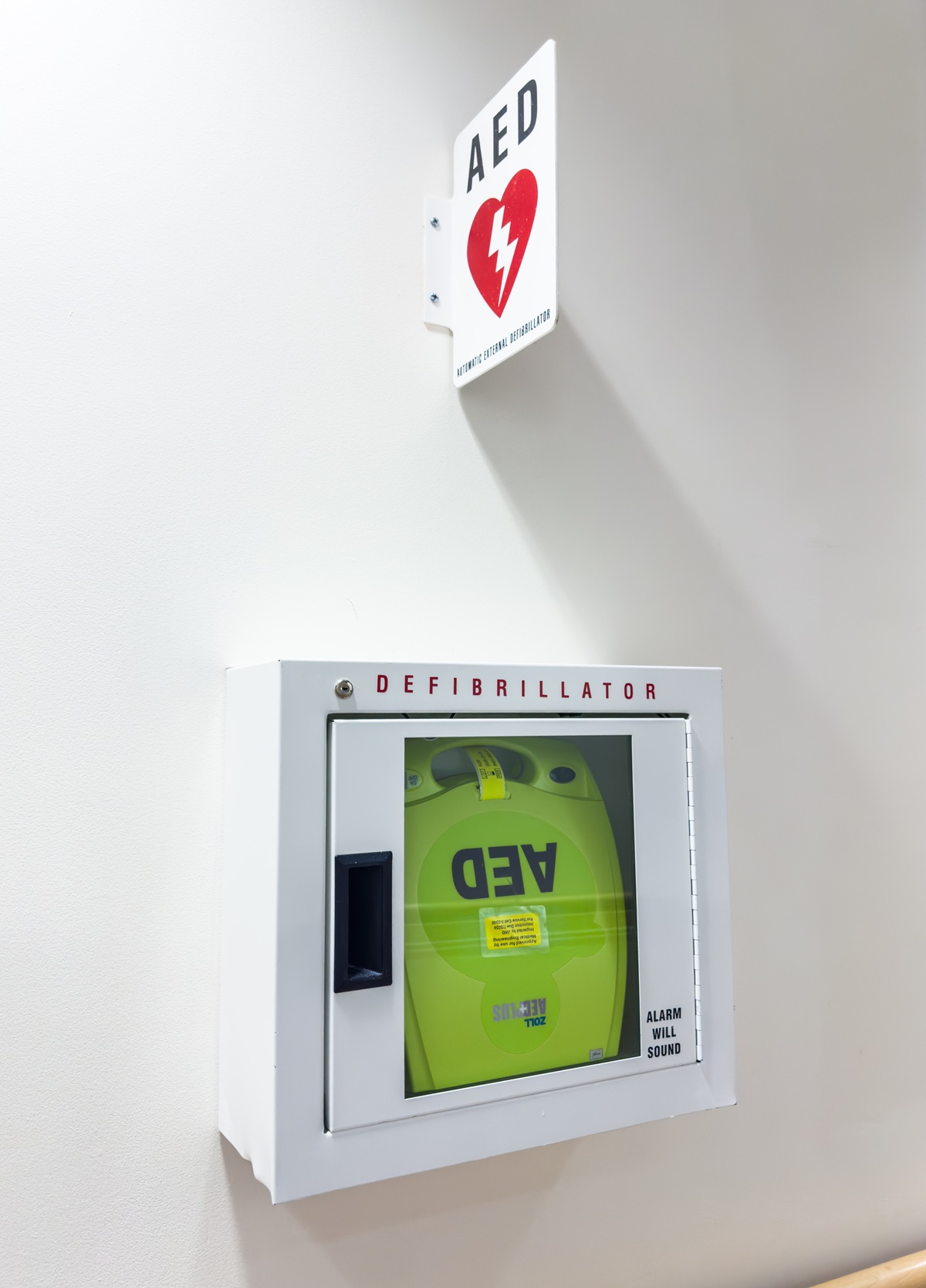 Automated external defibrillator stored in a wall cabinet beneath visible AED signage in a workplace hallway.